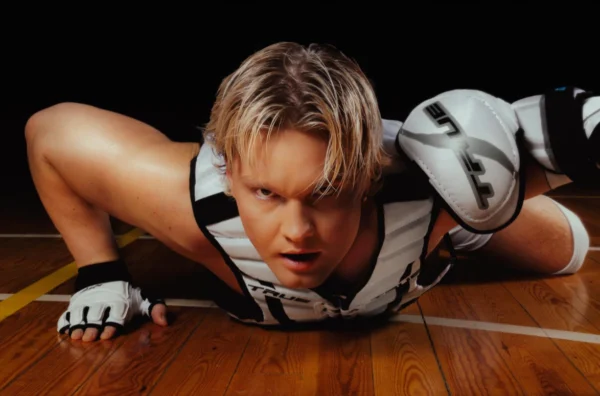 Benjamin in a dynamic pose, lying on the floor at the school gymnasium with his legs bent and head raised. The Benjamin has short blonde hair and wears a white and black sleeveless top with a thick black stripe around the armholes, paired with white and black shorts featuring a similar stripe at the waist. On his hands, he wear white fingerless gloves with black trim and ventilation holes at the fingertips and thumbs, secured by a strap that wraps over their forearm. Benjamin - Badabim