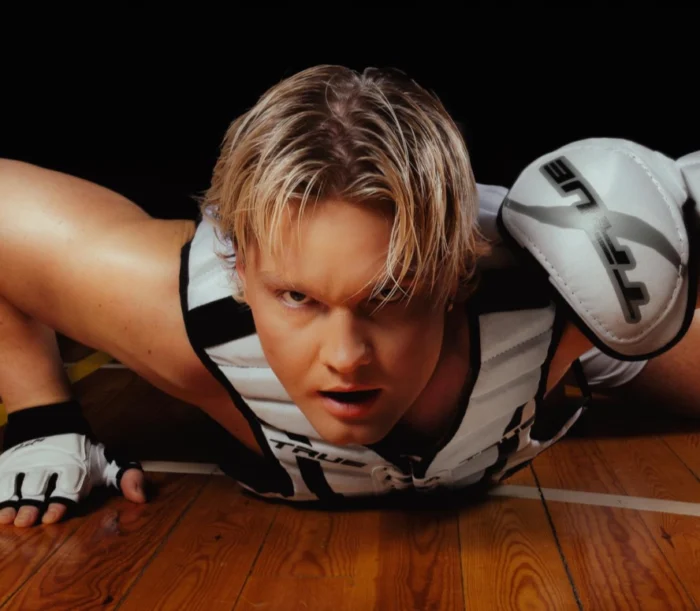 Benjamin in a dynamic pose, lying on the floor at the school gymnasium with his legs bent and head raised. The Benjamin has short blonde hair and wears a white and black sleeveless top with a thick black stripe around the armholes, paired with white and black shorts featuring a similar stripe at the waist. On his hands, he wear white fingerless gloves with black trim and ventilation holes at the fingertips and thumbs, secured by a strap that wraps over their forearm. Benjamin - Badabim